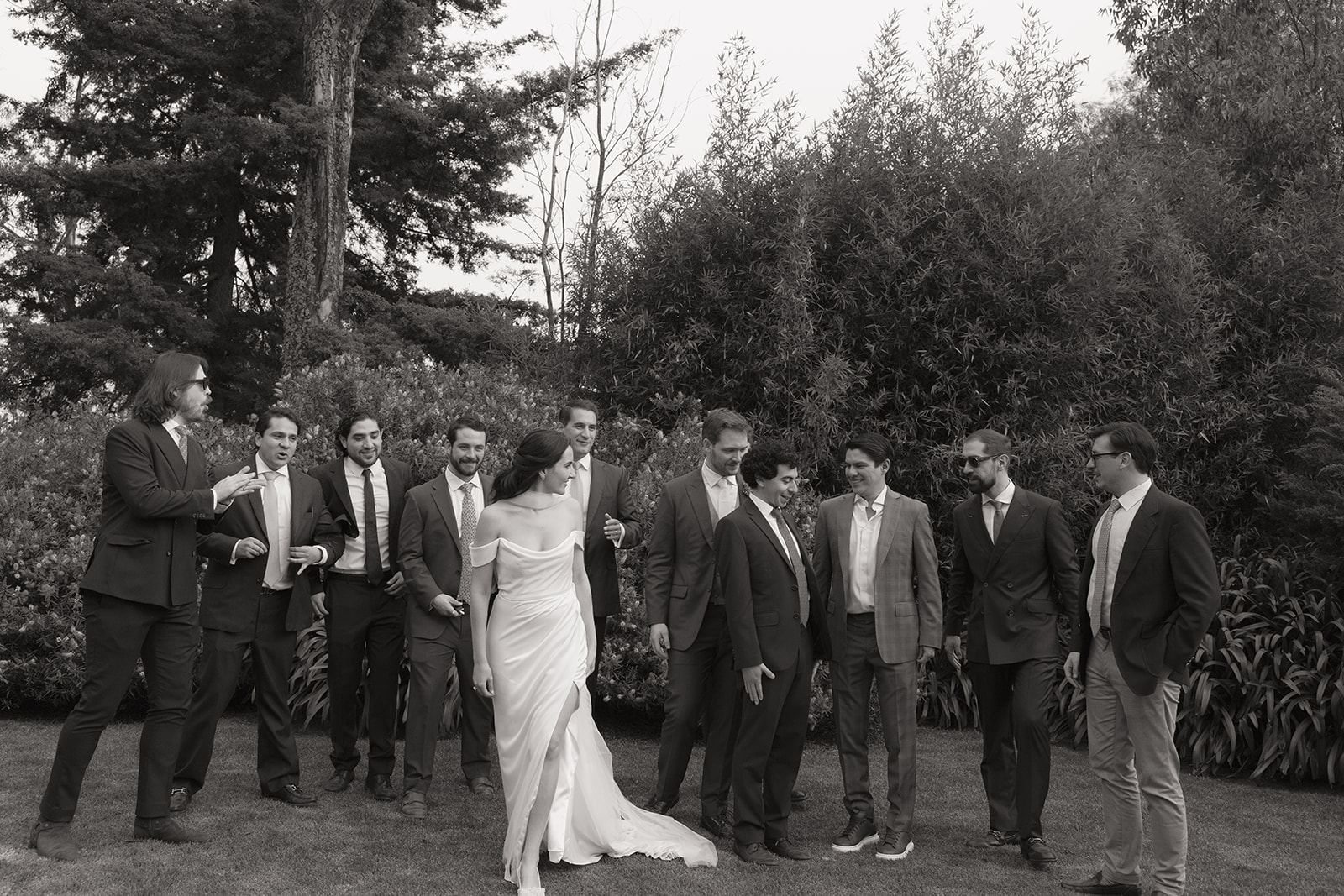 Bride laughing with groomsmen on a lawn surrounded by trees