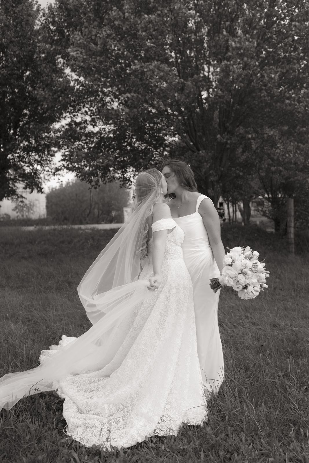 Two brides walking together across a grassy field under tall trees