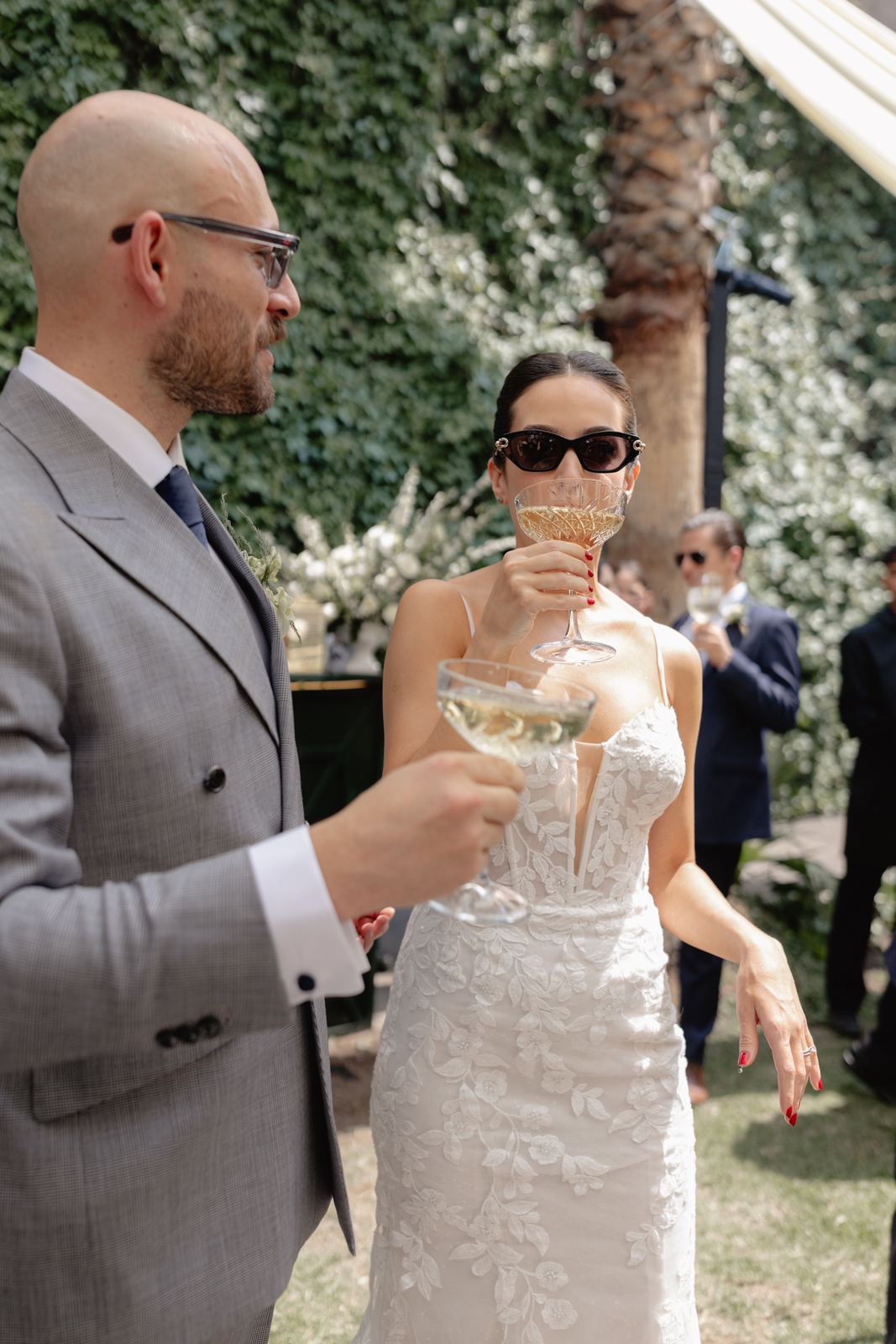 Bride in sunglasses sipping champagne beside the groom in a garden