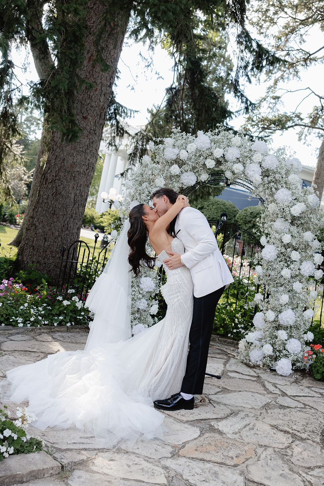 Couple kissing in front of a white floral arch on a stone patio