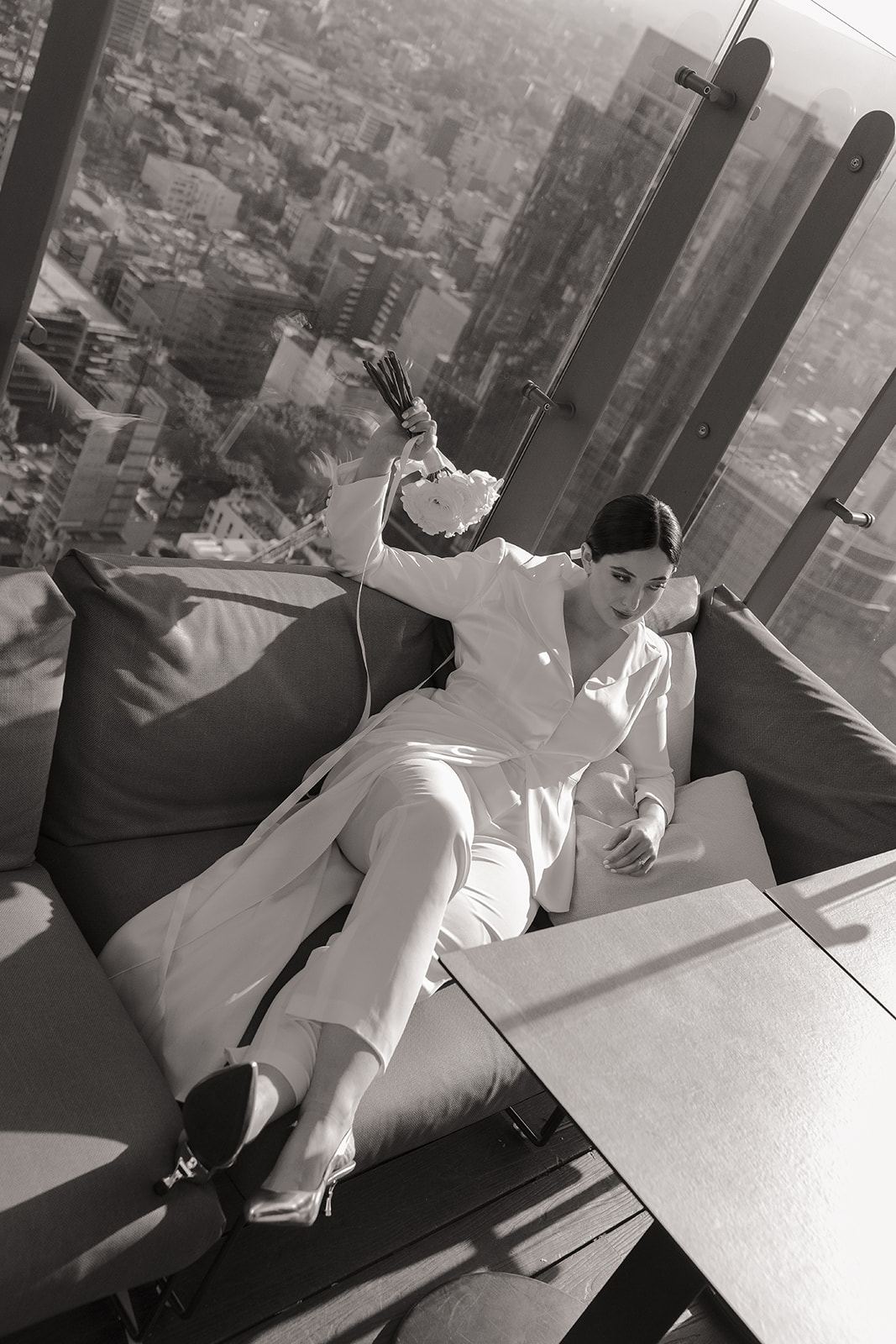 Bride in a white suit lounging by a high-rise window overlooking the city