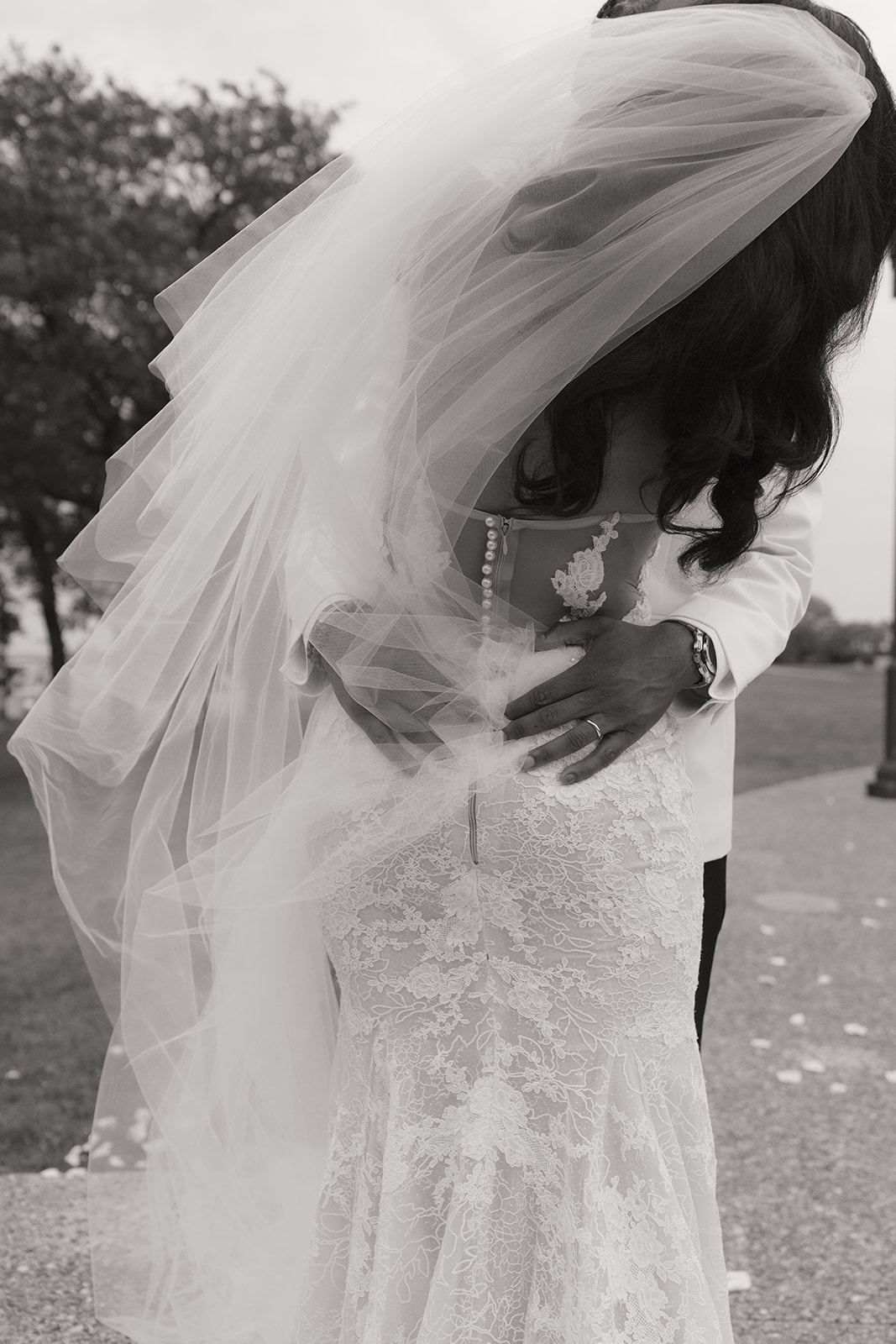 Groom's hand resting on the bride's lace back as her veil drifts