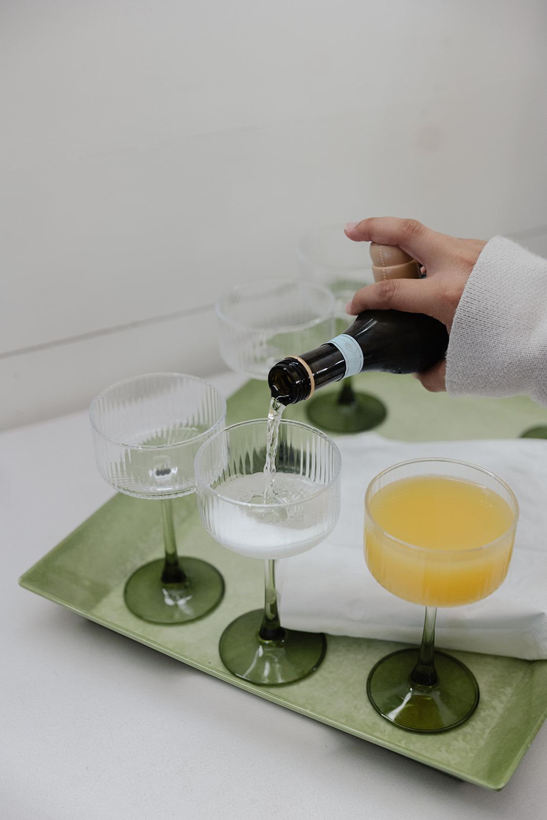 Pouring sparkling wine into coupes beside a mimosa on a green tray