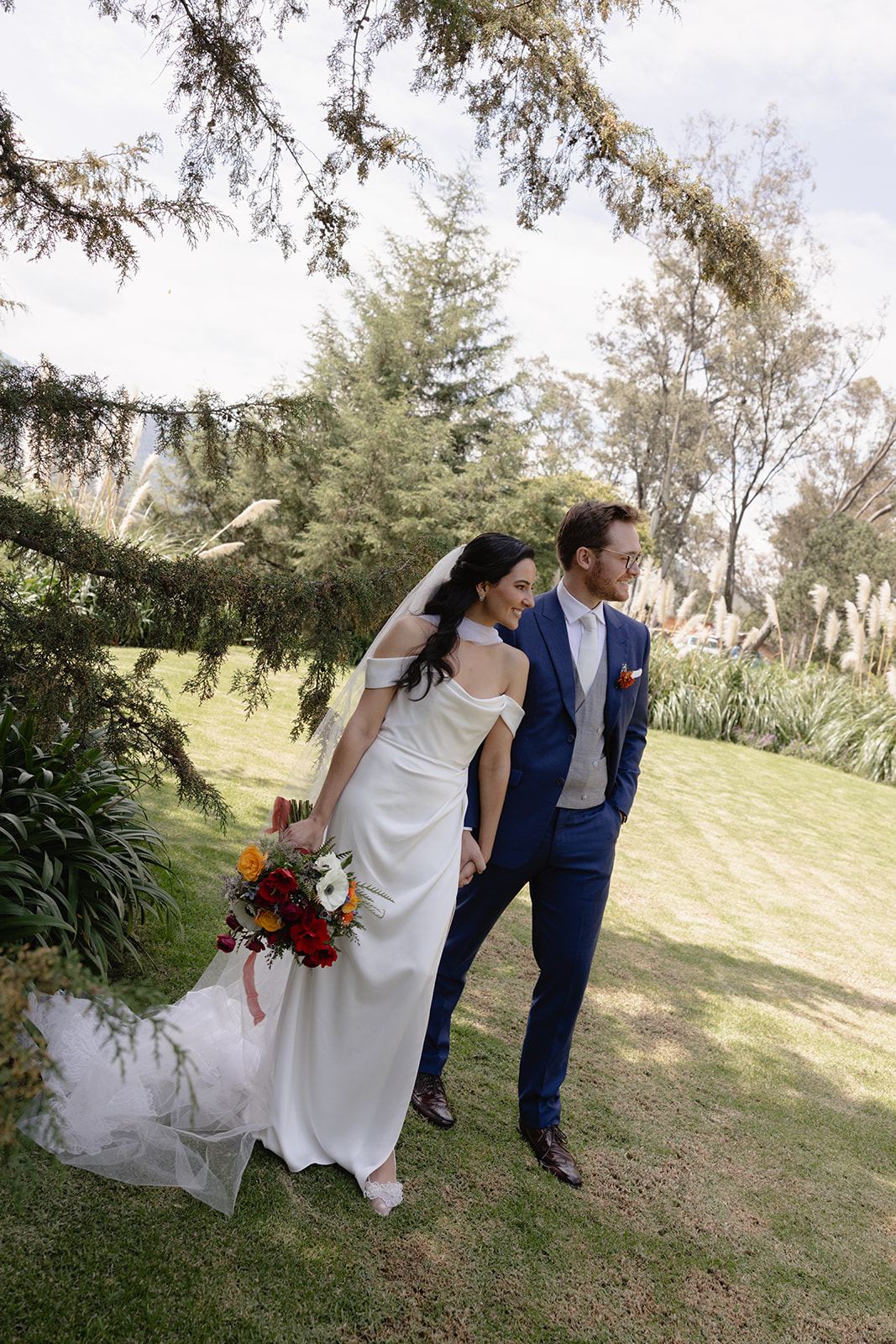 Couple strolling across a sunny lawn, the bride holding a red bouquet