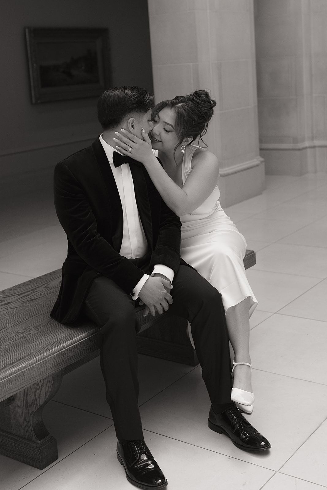Bride kissing the groom's cheek on a stone bench in a museum gallery