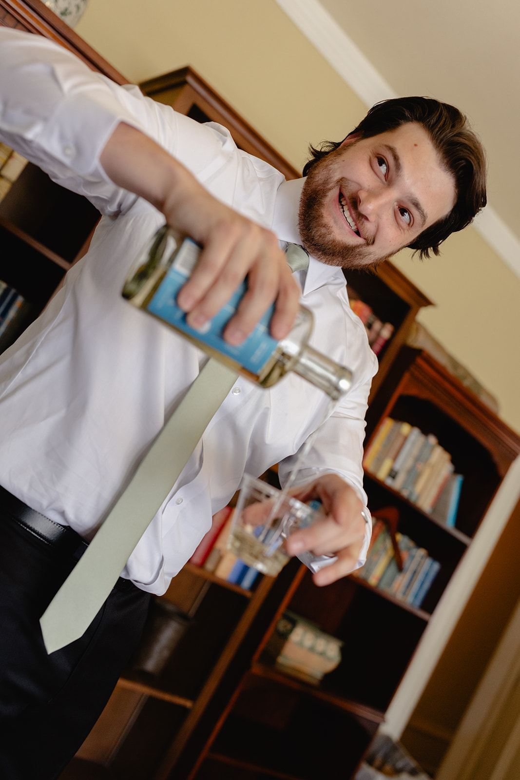Smiling man in a loose tie pouring drinks beside a bookcase